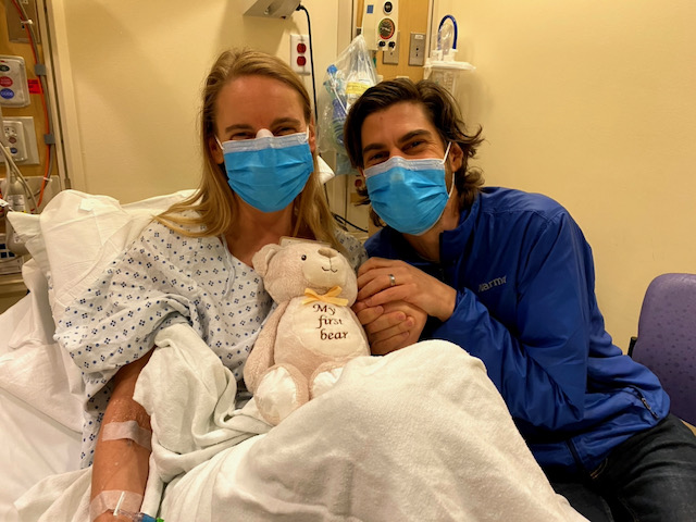 A white man and woman sit, masked, in a hospital room with a teddy bear on the woman's lap.